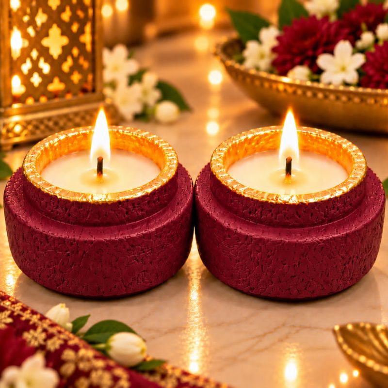 Two maroon textured candle holders with gold rims, lit, on a festive marble table